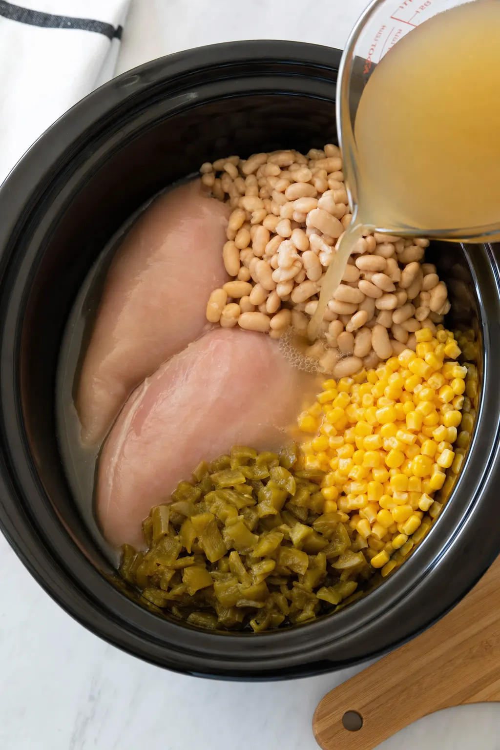 chicken beans corn and green chiles being loaded into slow cooker for white chicken chili